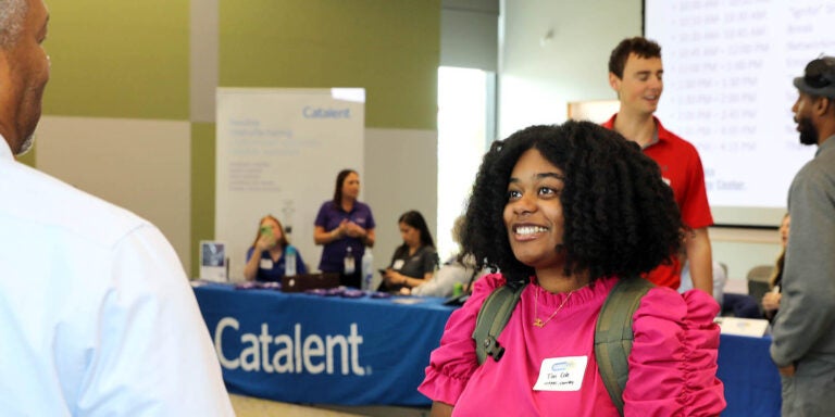 A woman in a red shirt smiles as she speaks with a man in a blue shirt as people stand in the background of an open room with table displays.