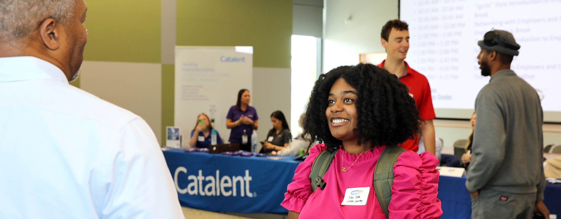 A woman in a red shirt smiles as she speaks with a man in a blue shirt as people stand in the background of an open room with table displays.