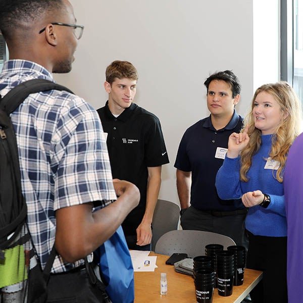 A woman at far right in a blue shirt stands next to two other men in black shirts as they talk to a taller person with his back to the camera.