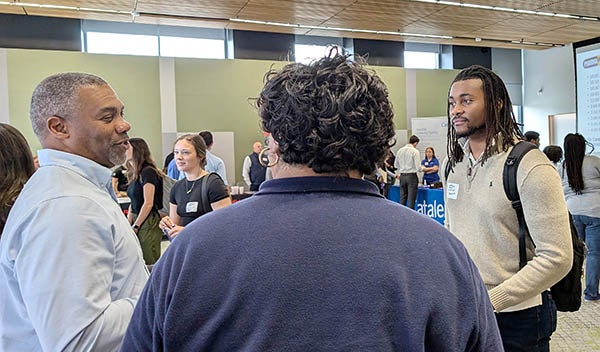 A man in a brown sweater at right stands and talks to two other men, one with his back to the camera and another on the left wearing a blue shirt.